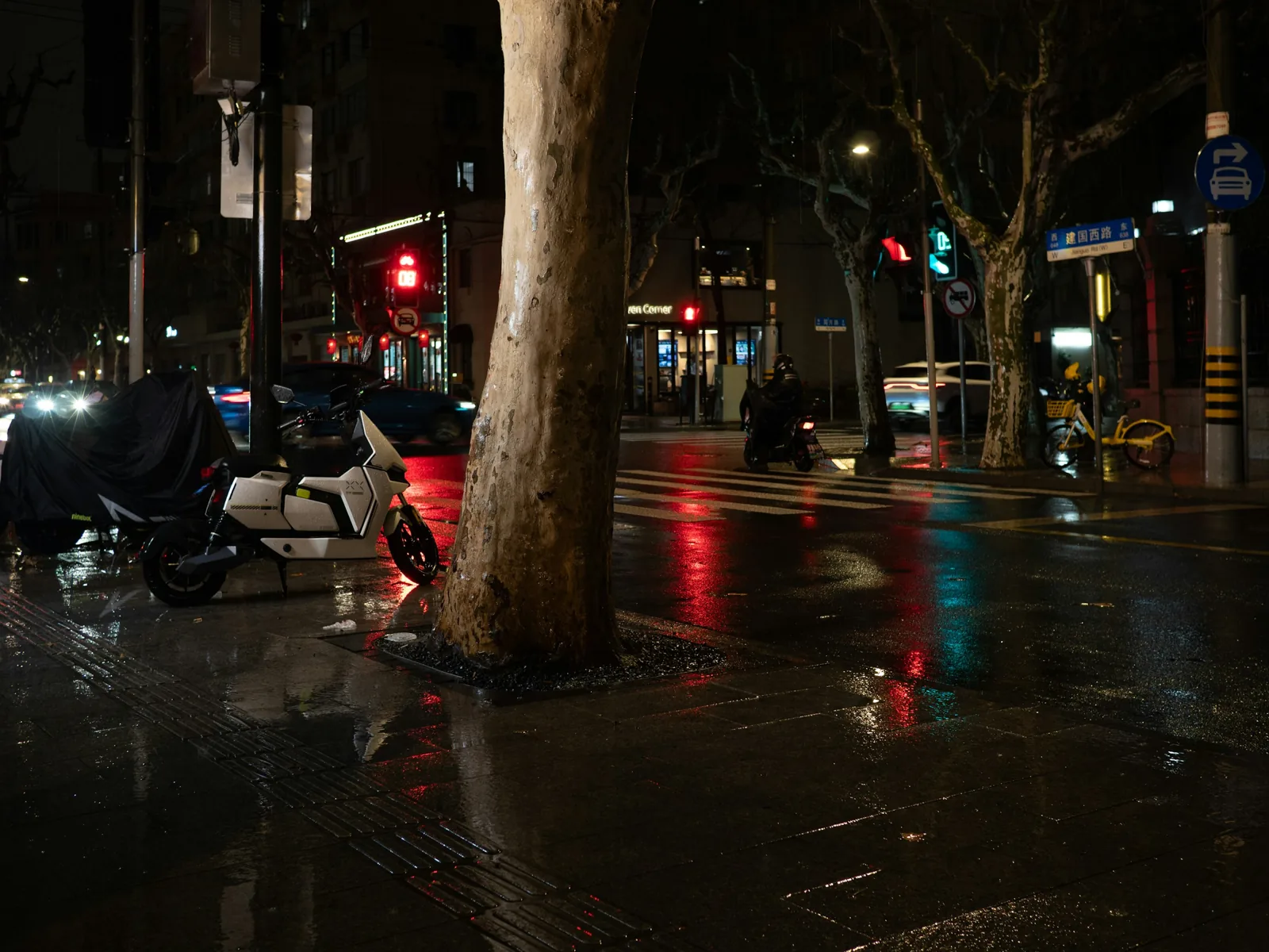 下過雨的夜晚街頭，濕亮的人行道上停著一台白色機車和一台蓋著車罩的車，遠處路口有紅綠燈與一位騎士